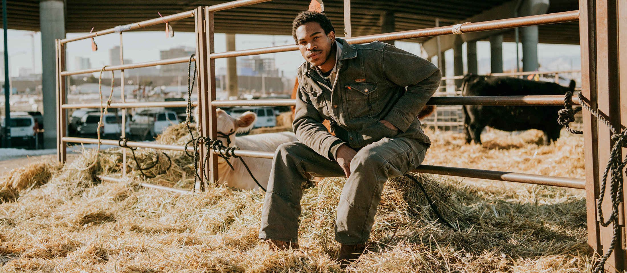 Man sits against fence wearing coveralls and posing in a cattle tie out with hay.